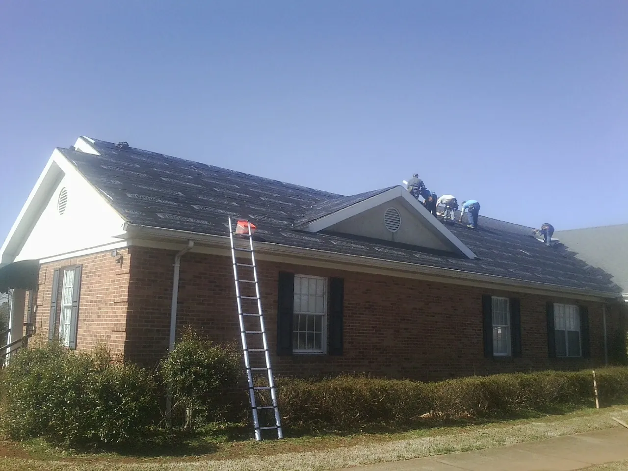 Crew tearing shingles off a brick ranch roof with a ladder set — Emergency Roof Repair work by our crew in Elk Park