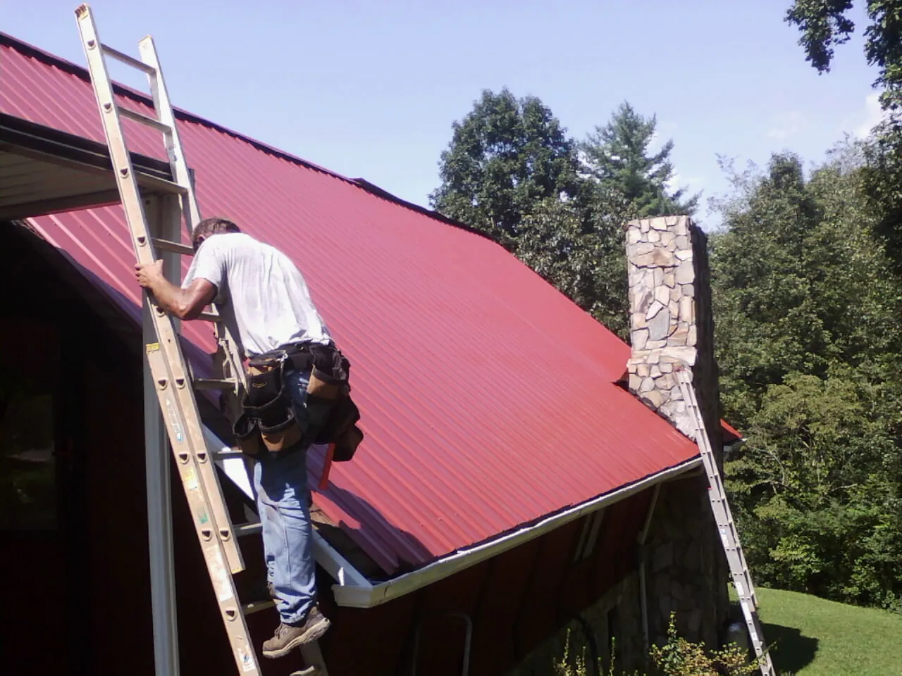 Roofer on a ladder beside a red metal roof and stone chimney — Soffit & Fascia Repair by Roofing Brothers in Elk Park